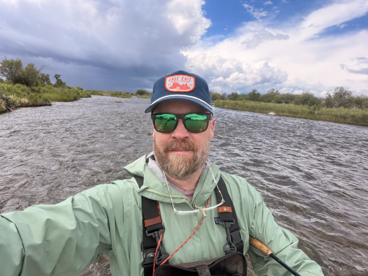 A man wearing a light green waterproof jacket, a navy rope Last Exit Goods hat, and green-tinted sunglasses takes a selfie with fly rod in a river under a cloudy sky.