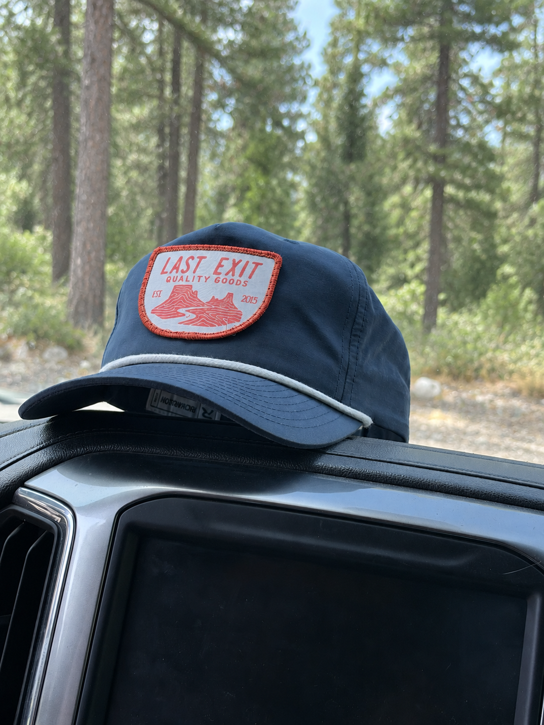 Side profile of the navy rope hat showing mesh flip-up stay, resting on the dashboard of a truck with a forest in the background.